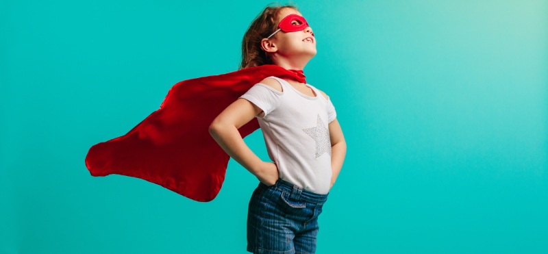 Child dressed up as a superhero with a red cape flowing against a turquoise background.