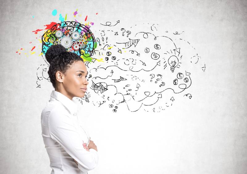 Woman thinking while standing with her arms folded, with a colorful brain image behind her on the wall displaying her thoughts.